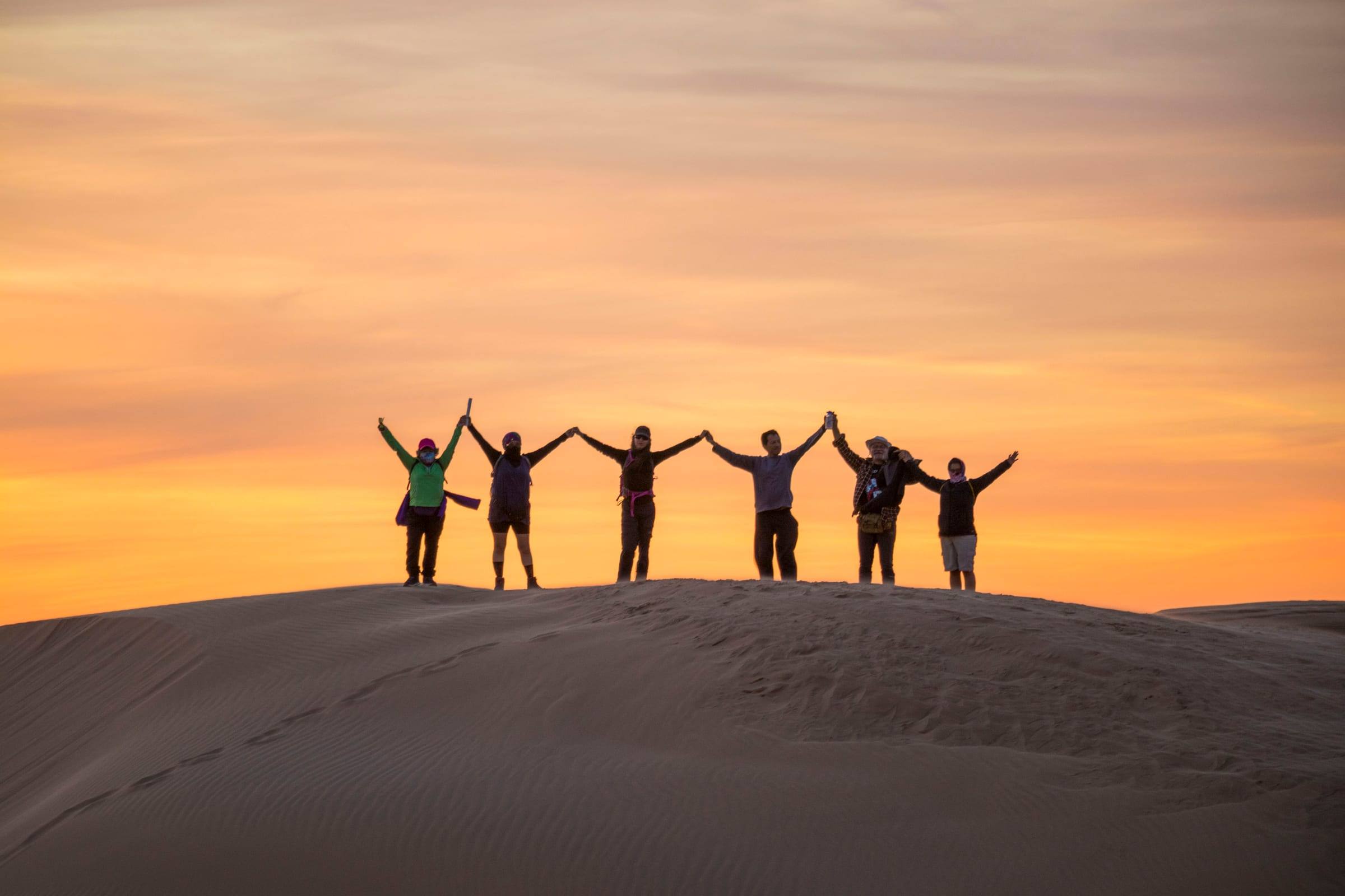 Grupo de viajeros con los brazos arriba en la cresta de una duna al atardecer